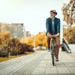 Ein Mann, der einen Helm, eine Brille und einen Blazer trägt, fährt an einem Herbsttag mit dem Fahrrad auf einem gepflasterten Weg durch einen Park, lächelt und blickt zur Seite.