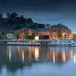 Ein ruhiger Fluss spiegelt die beleuchteten historischen Backsteingebäude und eine helle runde Straßenlaterne in der Abenddämmerung wider, mit einer Festung auf einem Hügel und dunklen Bäumen im Hintergrund. Ein angedocktes Boot ist entlang der Uferpromenade zu sehen.