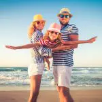 Eine lächelnde dreiköpfige Familie mit gestreiften Hemden, Hüten und Sonnenbrillen posiert spielerisch an einem sonnigen Strand, wobei die Eltern ihr Kind festhalten, während es die Arme wie ein Flugzeug ausstreckt. Im Hintergrund sind Wellen und blauer Himmel zu sehen.