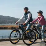 Ein Mann und ein junges Mädchen mit Helmen fahren an einem sonnigen Tag mit dem Fahrrad einen Uferweg entlang, mit Hügeln und Bäumen im Hintergrund.