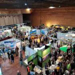 Eine große überdachte Ausstellungshalle ist voller Menschen, die sich zwischen den verschiedenen Ständen und Auslagen mit Bannern und Informationsständen bewegen. Es herrscht eine geschäftige und lebhafte Atmosphäre, in der die Besucher mit den Ausstellern ins Gespräch kommen.