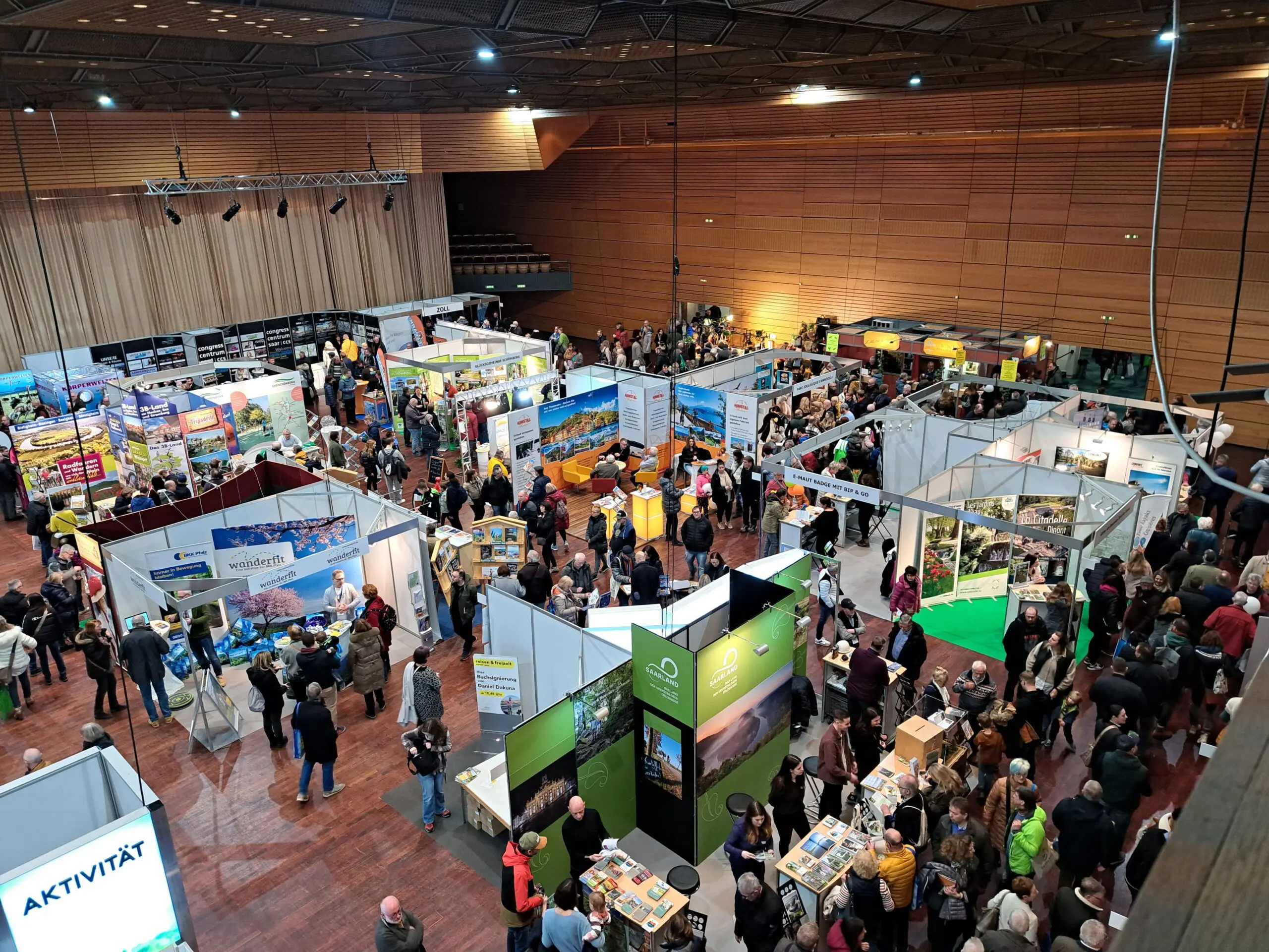 Luftaufnahme einer belebten Indoor-Ausstellung oder -Messe mit zahlreichen Ständen mit Bannern und Displays. Viele Menschen gehen umher, unterhalten sich und erkunden die verschiedenen Stände auf einem polierten Holzboden.