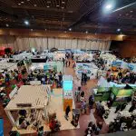 Eine belebte überdachte Ausstellungshalle, in der viele Menschen die verschiedenen Stände und Ausstellungen besuchen, mit Bannern, Tischen und Informationsständen, die über den großen, gut beleuchteten Raum verteilt sind.