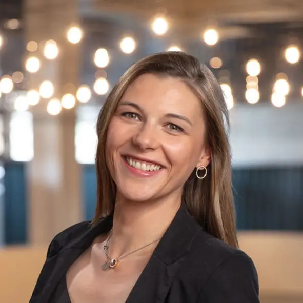 Eine Frau mit glattem braunem Haar, die einen schwarzen Blazer und silberne Ohrringe trägt, lächelt in die Kamera. Im Hintergrund sind verschwommene Hängelampen zu sehen, die eine warme und einladende Atmosphäre schaffen.