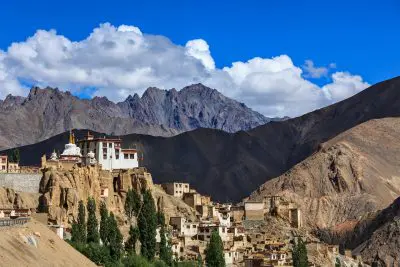 Ein Bergdorf im Himalaya mit traditionellen Häusern an felsigen Hängen, ein weißes Kloster mit einer goldenen Stupa, grüne Bäume und schroffe Gipfel unter einem blauen Himmel mit weißen Wolken.