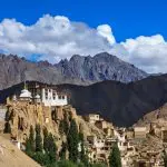 Ein Bergklosterkomplex mit weißen Gebäuden thront auf felsigen Klippen, umgeben von einem kleinen Dorf und hohen grünen Bäumen, mit schroffen Bergen und einem strahlend blauen Himmel mit weißen Wolken im Hintergrund.