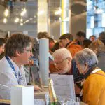 Ein Mann mit Namensschild spricht in einer belebten Halle mit einem älteren Ehepaar über einen Tresen hinweg. Im Hintergrund sind Menschen versammelt, und auf dem Tresen sind Dokumente und eine Sektflasche zu sehen.