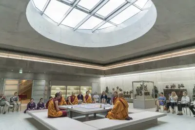 Vier buddhistische Mönche sitzen um einen Tisch herum und gestalten ein Sandmandala in einem modernen, runden Raum mit einem großen Oberlicht. Die Besucher sitzen ruhig um den Raum herum und beobachten den komplizierten Prozess und die umliegenden Ausstellungen buddhistischer Kunst.