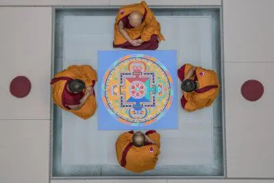 Vier buddhistische Mönche in orangefarbenen und roten Gewändern sitzen um ein farbenfrohes Sandmandala, das von oben betrachtet wird. Jeder Mönch ist einer anderen Seite des Mandalas zugewandt, daneben liegen runde rote Kissen auf einem hellen Boden.