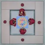 Vier Mönche in roten Roben sitzen um ein kompliziertes, farbenfrohes Mandala, das von oben betrachtet wird. Sie befinden sich in einem quadratischen, mit Kissen ausgekleideten Bereich mit vier runden roten Kissen an den Ecken.