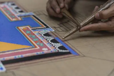 Nahaufnahme von Händen, die ein kompliziertes Sandmandala mit farbigem Sand auf einer hölzernen Oberfläche erstellen, wobei traditionelle Metallwerkzeuge verwendet werden, um den Sand sorgfältig in detaillierten Mustern und geometrischen Formen zu platzieren.