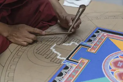 Eine Person verwendet traditionelle Werkzeuge, um sorgfältig ein farbenfrohes Sandmandala auf einem detaillierten kreisförmigen Umriss zu erstellen, wobei komplizierte Muster und leuchtende Farben aus einem Abschnitt herausragen.
