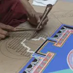 Eine Person verwendet traditionelle Werkzeuge, um sorgfältig ein kompliziertes Sandmandala auf einem detaillierten Umriss zu erstellen, wobei leuchtend blaue, weiße und mehrfarbige Abschnitte sichtbar sind.
