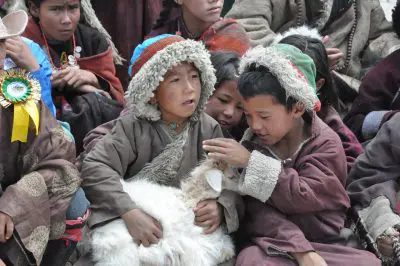 Eine Gruppe von Kindern in warmer, traditioneller Kleidung sitzt zusammen. Zwei Jungen im Vordergrund halten und streicheln sanft eine weiße Ziege, und alle wirken beschäftigt und zufrieden.