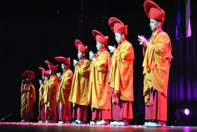 Eine Reihe von Mönchen in orangefarbenen und roten Roben mit hohen roten Hüten steht auf der Bühne, die Hände zum Gebet gefaltet, beleuchtet von der Bühnenbeleuchtung vor einem dunklen Hintergrund.