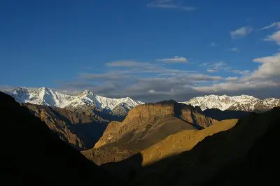 Berglandschaft mit schneebedeckten Gipfeln unter blauem Himmel, felsigen Hügeln im Vordergrund und dramatischem Sonnenlicht, das Schatten auf das Gelände wirft.