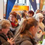 Eine überfüllte Veranstaltung in einer Halle mit Menschen verschiedenen Alters, die teilweise Papiere und Broschüren in der Hand halten, sich unterhalten und um Ausstellungsstände mit Bannern und Displays im Hintergrund herumgehen.