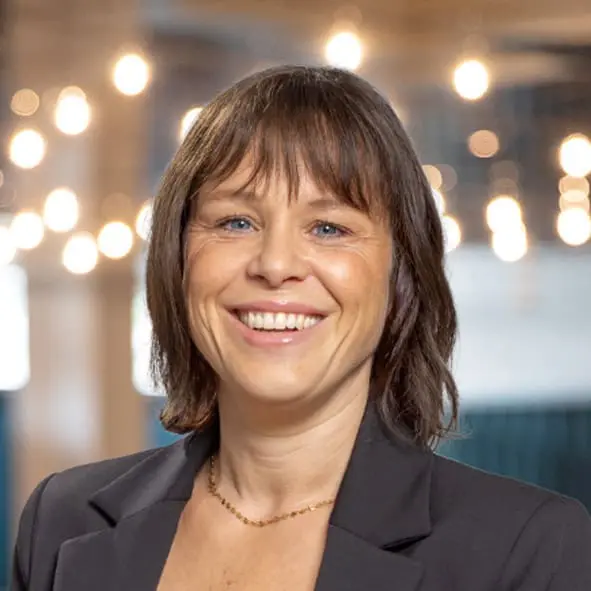 Eine Frau mit kurzem braunem Haar und Pony, die einen schwarzen Blazer und eine goldene Halskette trägt, lächelt vor einem verschwommenen Hintergrund mit Hängelampen.