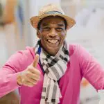 Ein lächelnder Mann, der einen Strohhut, einen rosa Pullover und einen schwarz-weiß gestreiften Schal trägt, zeigt den Daumen nach oben. Der Hintergrund ist sanft verschwommen.