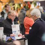 Ein älterer Mann hält eine Broschüre in der Hand und spricht mit einer Frau auf einer Indoor-Veranstaltung oder Messe. Im unscharfen Hintergrund sind weitere Personen zu sehen, was auf ein geschäftiges, öffentliches Umfeld hindeutet.