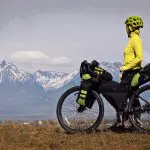 Ein Radfahrer mit gelbem Helm und gelber Jacke steht mit einem voll beladenen Fahrrad da und blickt auf ferne schneebedeckte Berge unter einem bewölkten Himmel.