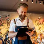 Eine lächelnde Person mit Bart, die eine Latzhose und ein weißes T-Shirt trägt, schreibt auf einem Klemmbrett in einem Fahrradgeschäft, in dem verschiedene Fahrräder an der Wand hinter ihnen ausgestellt sind.