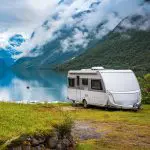 Ein weißer Wohnwagen steht auf einer Wiese am Ufer eines ruhigen Sees, umgeben von grünen Bergen mit schneebedeckten Gipfeln und dunstigen Wolken. Die friedliche Szene spiegelt sich im Wasser unter einem wolkenverhangenen Himmel.