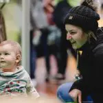 Eine lächelnde Frau mit schwarzer Wintermütze hockt neben einem Baby im Kapuzenpulli, im Hintergrund ist ein unscharfes Poster einer Wildkatze zu sehen. Beide scheinen drinnen bei einer Veranstaltung mit anderen Menschen zu sein.