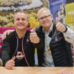 Zwei lächelnde Männer sitzen nebeneinander an einem Tisch und machen Daumen-nach-oben-Gesten. Sie tragen beide Jacken und bunte Medaillen um den Hals. Hinter ihnen ist ein bunter, verschwommener Hintergrund mit Text und Bildern zu sehen.