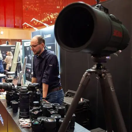 Ein Mann steht hinter einem mit Kameraobjektiven gefüllten Ausstellungstisch bei einer Veranstaltung. Im Vordergrund ist ein großes Sigma-Teleobjektiv auf einem Stativ montiert. Im Hintergrund sind weitere Personen und Stände zu sehen.