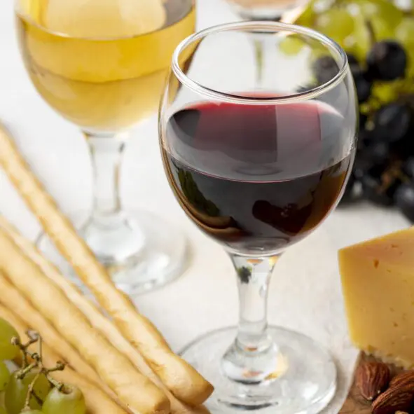 Zwei Weingläser, eines mit Rotwein und eines mit Weißwein, umgeben von Brotstangen, grünen Weintrauben, einer Käsescheibe und einigen Nüssen auf einer weißen Fläche.