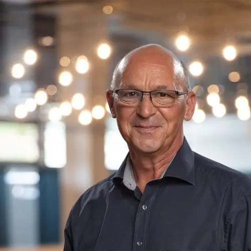 Ein lächelnder älterer Mann mit Brille und dunklem Hemd steht in einem Haus, in dem die warmen Hängelampen im Hintergrund verschwimmen.