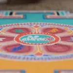 Nahaufnahme eines farbenfrohen Sandmandalas mit komplizierten kreisförmigen Mustern, einschließlich roter, grüner, oranger und blauer Formen, die auf einer flachen Oberfläche entstanden sind. Der Schwerpunkt liegt auf dem detaillierten Muster in der Mitte.