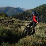 Eine Person, die einen blauen Helm und ein rotes Hemd trägt, fährt mit einem Mountainbike auf einem holprigen Weg, der von grünen Sträuchern umgeben ist, mit bewaldeten Hügeln und fernen Bergen im Hintergrund unter einem klaren Himmel.