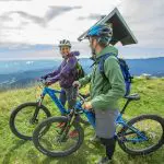 Zwei Personen mit Helmen und Outdoor-Ausrüstung stehen mit ihren Mountainbikes auf einer grasbewachsenen Hügelkuppe und lächeln sich an, während im Hintergrund die malerischen Berge und eine kleine Holzhütte zu sehen sind.