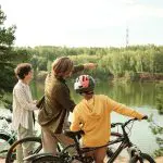 Drei Personen mit Fahrrädern stehen an einem Fluss. Ein Erwachsener zeigt über das Wasser, während ein Kind mit Helm und eine weitere Person in die gleiche Richtung schauen. Bäume und Grünflächen umgeben den Fluss unter einem klaren Himmel.