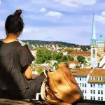 Eine Frau mit einem Dutt sitzt auf einem Felsvorsprung und blickt auf eine Stadtlandschaft, neben sich einen großen braunen Rucksack. Im Hintergrund sind Dächer, grüne Bäume und eine Kirche mit einem hohen blauen Kirchturm zu sehen.