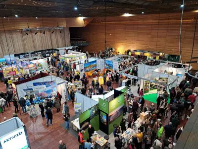 Eine große überdachte Kongresshalle, in der viele Menschen zwischen zahlreichen Ausstellungsständen mit Bannern und Displays verschiedener Unternehmen und Organisationen umherlaufen; der Raum ist hell erleuchtet und geschäftig.