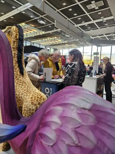 Drei Personen stehen an einem überdachten Stand und unterhalten sich. Im Vordergrund befinden sich große ausgestopfte Tierfiguren, eine Giraffe und ein lila Vogel. Darüber ist eine helle Deckenbeleuchtung zu sehen.
