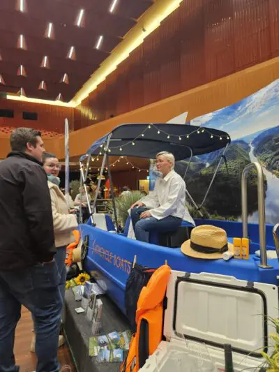 Drei Personen unterhalten sich bei einer Indoor-Veranstaltung. Eine Frau sitzt in einem blauen Tretboot mit Lichterketten und Flusslandschaft, während zwei andere in der Nähe stehen. Rund um den Stand liegen Schwimmwesten, Hüte und Broschüren aus.