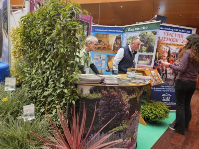 An einem Messestand stehen zwei Männer hinter einem mit Gartenbildern dekorierten Tresen, umgeben von üppigen Pflanzen. Eine Frau steht davor und sieht sich Broschüren an. Im Hintergrund sind bunte Banner zu sehen, die für die Villa Borg und eine Veranstaltung werben.