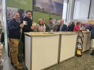 Fünf Personen stehen lächelnd hinter einem Ausstellungsstand mit Broschüren und Flyern in einem großen, hellen Raum. Im Hintergrund sind andere Besucher und Stände zu sehen.