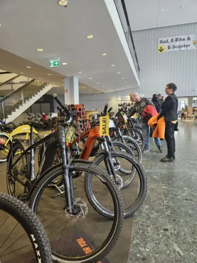 In der Halle ist eine Reihe von Fahrrädern ausgestellt, die von mehreren Personen begutachtet werden. Die Schilder darüber zeigen Abteilungen für Fahrräder, E-Bikes und E-Scooter an. Die Umgebung scheint ein Ausstellungsraum oder eine Ausstellungshalle zu sein.