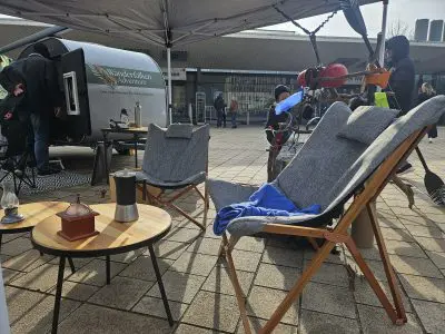 Zwei gepolsterte Campingstühle und ein kleiner runder Tisch mit Kaffeezubehör sind im Freien in der Nähe eines Anhängers aufgestellt. Menschen in Jacken stehen in der Nähe unter einem Zelt auf einem gepflasterten Platz.