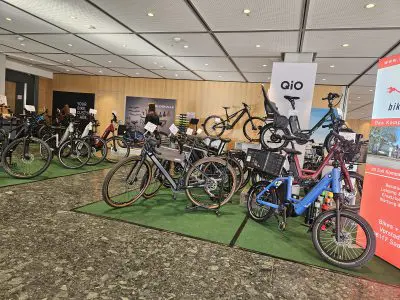 Eine Ausstellung verschiedener Fahrräder in einem Ausstellungsraum, mit E-Bikes und Lastenrädern auf grünen Matten. Im Hintergrund sind Banner und Plakate zu sehen, die für verschiedene Fahrradmarken und -modelle werben.