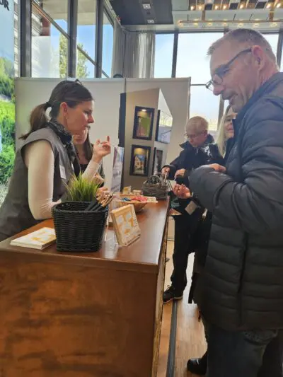 Eine Frau hinter einem Tresen spricht mit mehreren Personen bei einer Veranstaltung in einer Halle. Auf dem Tresen stehen eine kleine Pflanze, Broschüren und Snacks. An den Wänden im Hintergrund sind gerahmte Bilder ausgestellt.