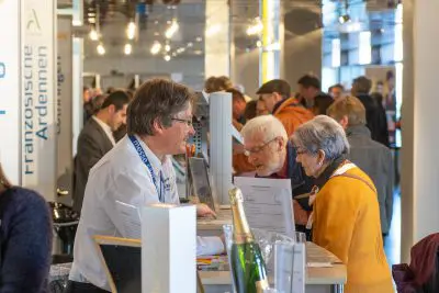 Ein Mann an einem Informationsstand lächelt und unterhält sich mit zwei älteren Erwachsenen, einem Mann und einer Frau, bei einer belebten Veranstaltung in einer Halle mit anderen Menschen im Hintergrund.