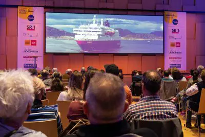 Ein Publikum sitzt in einem hölzernen Auditorium und schaut auf eine große Leinwand, auf der das Bild eines Kreuzfahrtschiffs auf dem Meer mit Bergen im Hintergrund zu sehen ist. An den Wänden sind verschiedene Sponsorenbanner zu sehen.