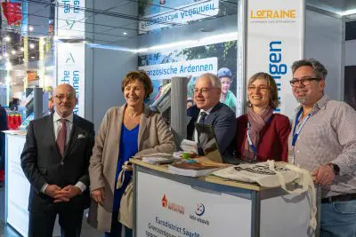 Fünf Erwachsene, drei Männer und zwei Frauen, stehen lächelnd an einem Messestand mit Broschüren, Tragetaschen und Schildern mit dem Hinweis auf den Eurodistrikt SaarMosel und Lothringen im Hintergrund.