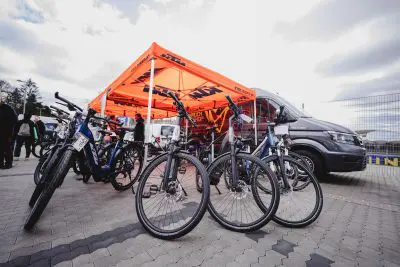 Mehrere Fahrräder sind unter einem orangefarbenen KTM-Zelt bei einer Freiluftveranstaltung ausgestellt, daneben parkt ein schwarzer Lieferwagen. Im Hintergrund sind an einem bewölkten Tag Menschen versammelt.
